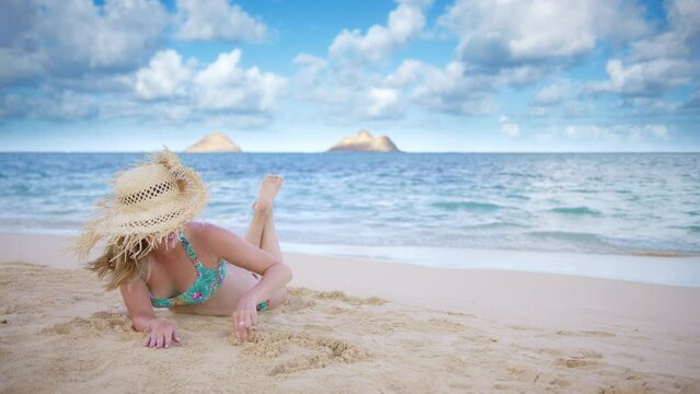 Smiling tourist in fringed straw hat drawing heart in golden sand on Oahu beach. Love and happiness concept. Female traveler sunbathing Lanikai beach Mokulua islands view. Young woman tanning on beach