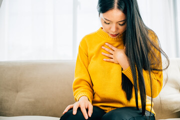 A woman from Asia suffers a heart attack on a sofa clutching her left chest in pain needing urgent medical aid. Depicting heartache discomfort and the urgency for emergency help.