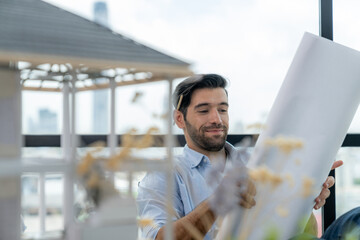 Smiling engineer looking project plan while design house model at modern office. Smart caucasian...