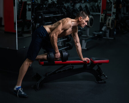 Shirtless man doing dumbbell row to waist on bench at gym. 