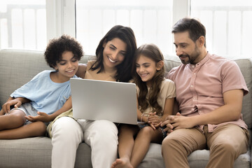 Cheerful Latin couple of parents and two cute kids talking on family group video call at laptop, resting on sofa together, holding computer, enjoying Internet communication, online entertainment