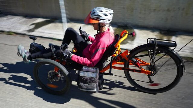 Rear side view of elderly senior woman riding recumbent tricycle e-bike on a path on a sunny day in a park.