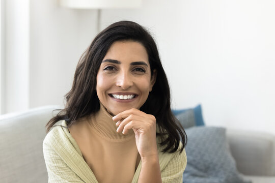 Cheerful beautiful middle aged Latin woman looking at camera with toothy smile, touching chin, showing white perfect healthy teeth, posing for beauty care portrait, sitting on home couch