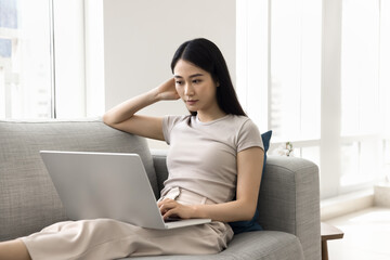 Focused young adult Chinese girl using online application on laptop, e-commerce service for shopping, Internet communication at home, typing, watching content, resting on sofa