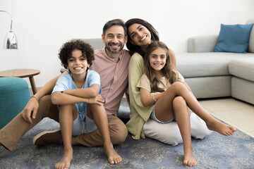 Happy Latin couple of parents and two cheerful kids sitting on carpeted floor at cozy home, looking...