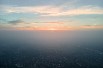 Aerial view from airplane window at high altitude of distant city covered with layer of thin misty smog and distant clouds in evening