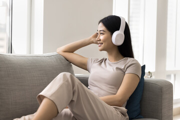 Obraz premium Positive relaxed young Asian music fan girl wearing wireless headphones, looking at window away, listening to songs from online playlist, musical Internet service, media application, relaxing at home