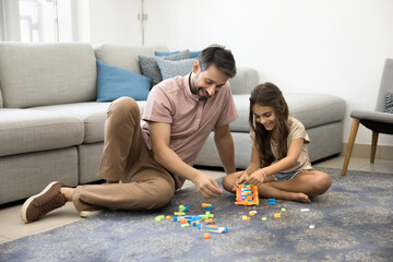 Happy daddy and cute little daughter girl constructing toy tower from building blocks, playing on warm carpeted floor, smiling, laughing, having fun on family playtime with child