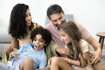 Fototapeta premium Cheerful loving Latin parents tickling excited kids, playing active games with preteen children on floor at home, having fun, laughing, enjoying family leisure, activity, parenthood