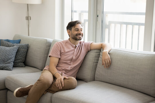 Positive thoughtful relaxed middle aged man sitting on comfortable couch, looking at window away, thinking, daydreaming, enjoying home leisure, silence, relaxation, comfort, smiling