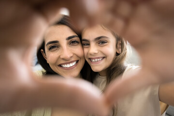 Happy little child girl and young mom looking at camera through finger heart, making hand frame, symbol of love, kindness, friendship, smiling, showing perfect white teeth, posing for family portrait