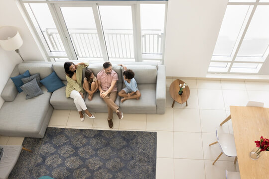 Young parents and two preteen sibling children sitting on couch in spacious apartment, talking, enjoying family home leisure in living room with sofa, carpet, dining table