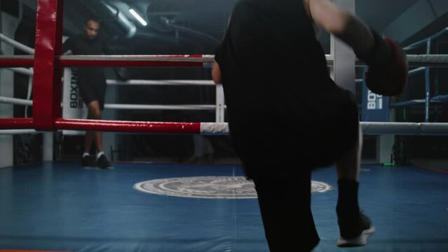 Teenager sits near boxing ring and puts on boxing gloves. Young boxer comes in boxing ring and starts practice punches with African American trainer. Athlete prepares to competition in boxing gym.