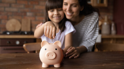 Saving money. Happy hispanic mother hug preteen daughter dropping coin into piggybank support child in wish to make savings teach economy financial planning. Soft focus on cute piggy bank toy on table