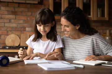 Mother assist in learning. Caring hispanic mom help preteen daughter in doing homework check written task correct mistake. Young female teacher explain grammar rule to little girl pupil at home lesson - obrazy, fototapety, plakaty