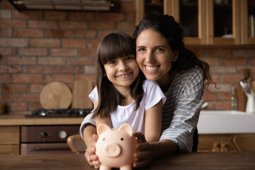 We choose wise economy. Portrait of happy little latin girl and single mom sit at kitchen table cuddle look at camera hold toy piggy bank. Thrifty mum parent and preteen kid make deposit save up money