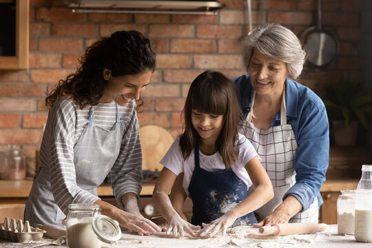 Hands in flour. Friendly intergenerational family of 3 diverse age females engaged in cooking short pastry biscuits. Young mom senior granny assist little girl stretch dough for pie on kitchen table