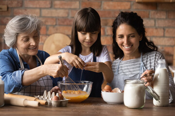 Funny family cooking. Three diverse age hispanic females relatives preteen girl young mom and elderly grandma mixing dough for cookies at kitchen. Happy little kid bake homemade cake with mum granny