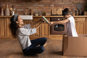 Ship is under attack. Overjoyed young single mom and small girl having fun play pirates at home kitchen. Laughing mexican nanny imitate battle on swords with kid capture defend boat made of carton box