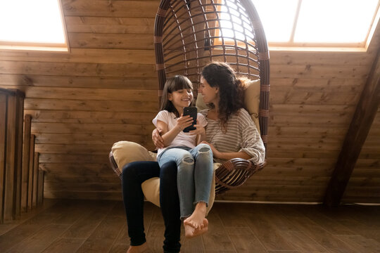 Smiling single latina mother relax in rattan chair swing at comfy attic hold preteen daughter on lap use smartphone. Friendly mom and child girl talk discuss news at social network play cellphone game