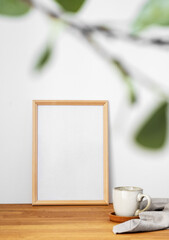 A frame with a blank canvas against a light wall and on a wooden tabletop with blurred green foliage and cup of tea