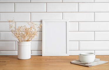 A  frame with a blank canvas against a white tile wall and on a wooden tabletop with vase and cup.