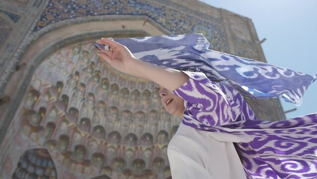 A young woman with long hair in an oriental robe and skullcap stands in the wind near an ancient mosque