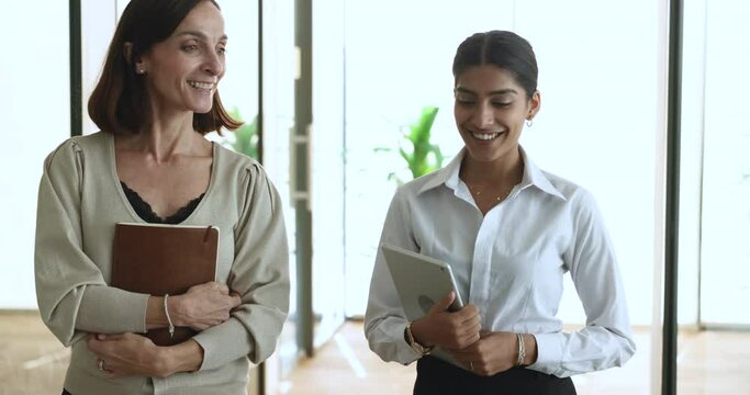 Two confident diverse younger and elder female colleagues walking in office hall, holding tablet, talking, listening, nodding, smiling, stopping at camera, using gadget for work discussion