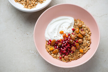 Obraz na płótnie Canvas Roseate bowl with granola, yogurt, pomegranate and nuts, horizontal shot on a light-beige stone background, elevated view