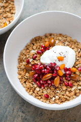 Granola bowl with yogurt, pomegranate seeds, nuts and honey, vertical shot on a beige granite surface, middle close-up, selective focus