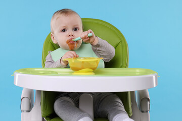 Cute little baby eating healthy food in high chair on light blue background