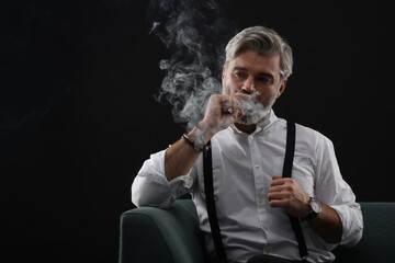 Bearded man smoking cigar on sofa against black background. Space for text
