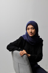 Serious Asian hijab woman in casual shirt sitting on chair, posing with various hand gesture. Isolated studio portrait on gray background.