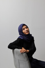 Serious Asian hijab woman in casual shirt sitting on chair, posing with various hand gesture. Isolated studio portrait on gray background.