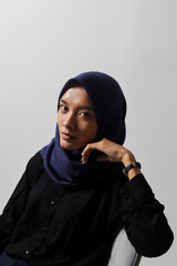 Serious Asian hijab woman in casual shirt sitting on chair, posing with various hand gesture. Isolated studio portrait on gray background.