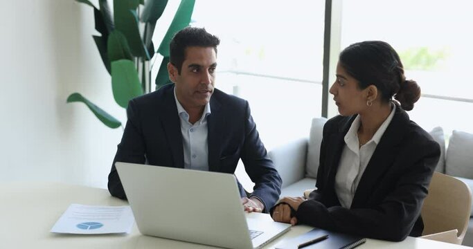 Two Indian business colleagues in formal suits talking at laptop computer, sitting at office workplace, discussing teamwork, cooperating on online project. Mentor training intern