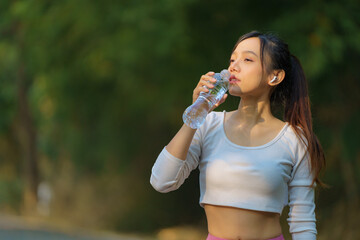 Close-up beautiful Asian woman drinking water from a bottle after running with a public park background, outdoor workout concept, drinking clear mineral water after jogging.