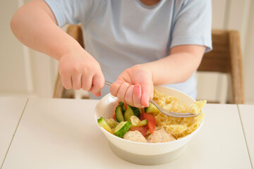 A child eats pasta with a fork while sitting at a table in a home kitchen. Little boy eating noodles with vegetables in a white bowl on the table. Toddler baby two years old at dinner