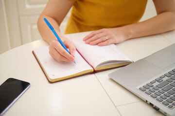 A woman with a laptop is writing with a pen in a notebook while sitting at a table in a home kitchen. An adult female businesswoman works from home, a remote office