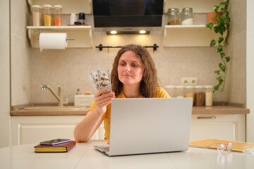 Fototapeta na wymiar A woman with a laptop holds money dollars at a table in a home kitchen. An adult female businesswoman works from home, a remote office