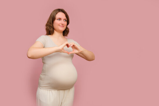 Pregnant woman shows heart with hands on studio pink background. Pregnancy in a woman with a belly with a hand showing love, copy space