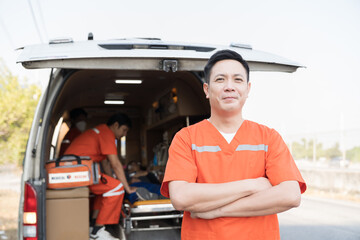 Asian male ambulance staff member standing with crossed arms. He is wearing ambulance uniform of paramedics during paramedics giving injured person first aid background © amorn