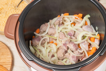 Fries meat with onions and carrots in a multicooker pan. Modern, bright multi cooker with products. Cooking pilaf with meat in multicooker. Top view of multi cooker with vegetables and meat.