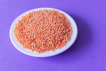 A pile of broken red lentils on a white plate on a purple background