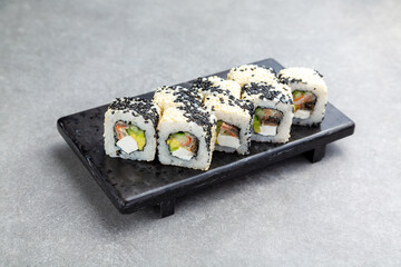 Japanese Sushi roll in black and white sesame with avocado, cheese and eel on a black stand, gray background