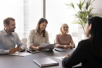 HR team of happy employers interviewing young Asian job candidate woman at meeting table, talking to applicant, reviewing paper resume, laughing, discussing vacancy - obrazy, fototapety, plakaty