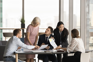 Multiethnic team of coworkers analyzing paper marketing reports at meeting table, brainstorming on sales result, reviewing documents together, discussing project strategy