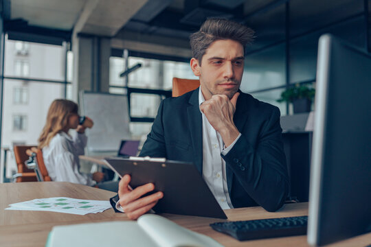 Male manager sitting at desk with clipboard and thinking about project while looks on computer - Powered by Adobe
