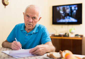 Elderly man writes a letter on a sheet of paper while sitting at a table in room