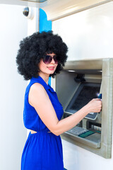 A woman in a blue dress wearing sunglasses withdraws cash from an ATM street.